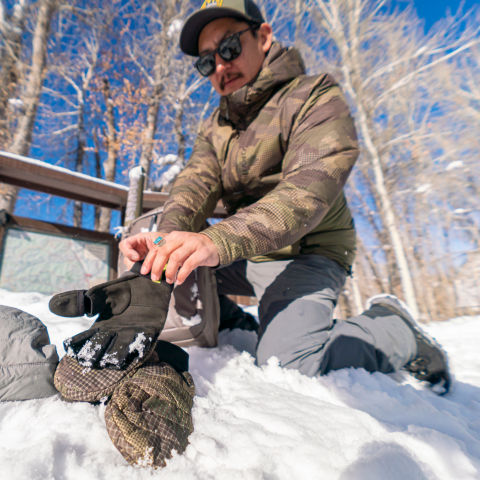 An angler kneeling in the snow puts on a pair of gloves that matches the camo of their hoodie